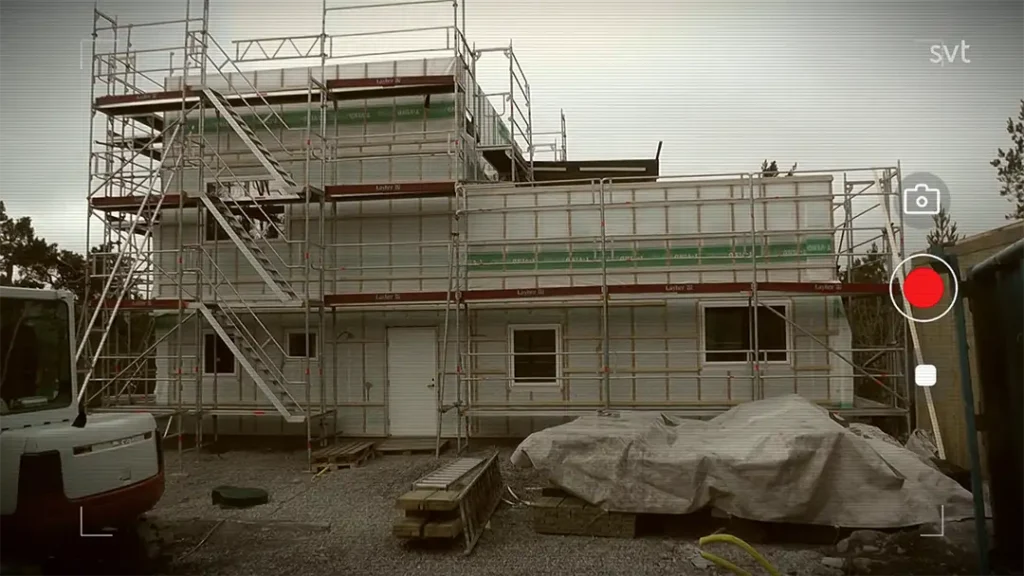 Stiligt - Älgö - Husdrömmar: En tvåvåningsbyggnad under uppförande med ställningar, täckta material och entreprenadutrustning synliga framför. Kamerainspelningsgränssnitt överlagrar bilden.