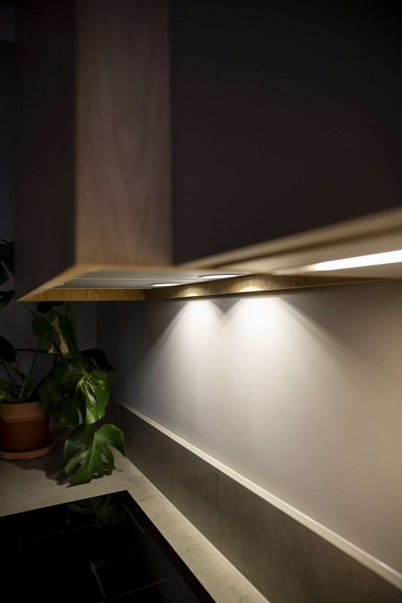 Stiligt - En köksbänk med en stiligt krukväxt och ett överskåp i trä med belysning under skåpet som elegant lyser upp baksidan och en svart spishäll.