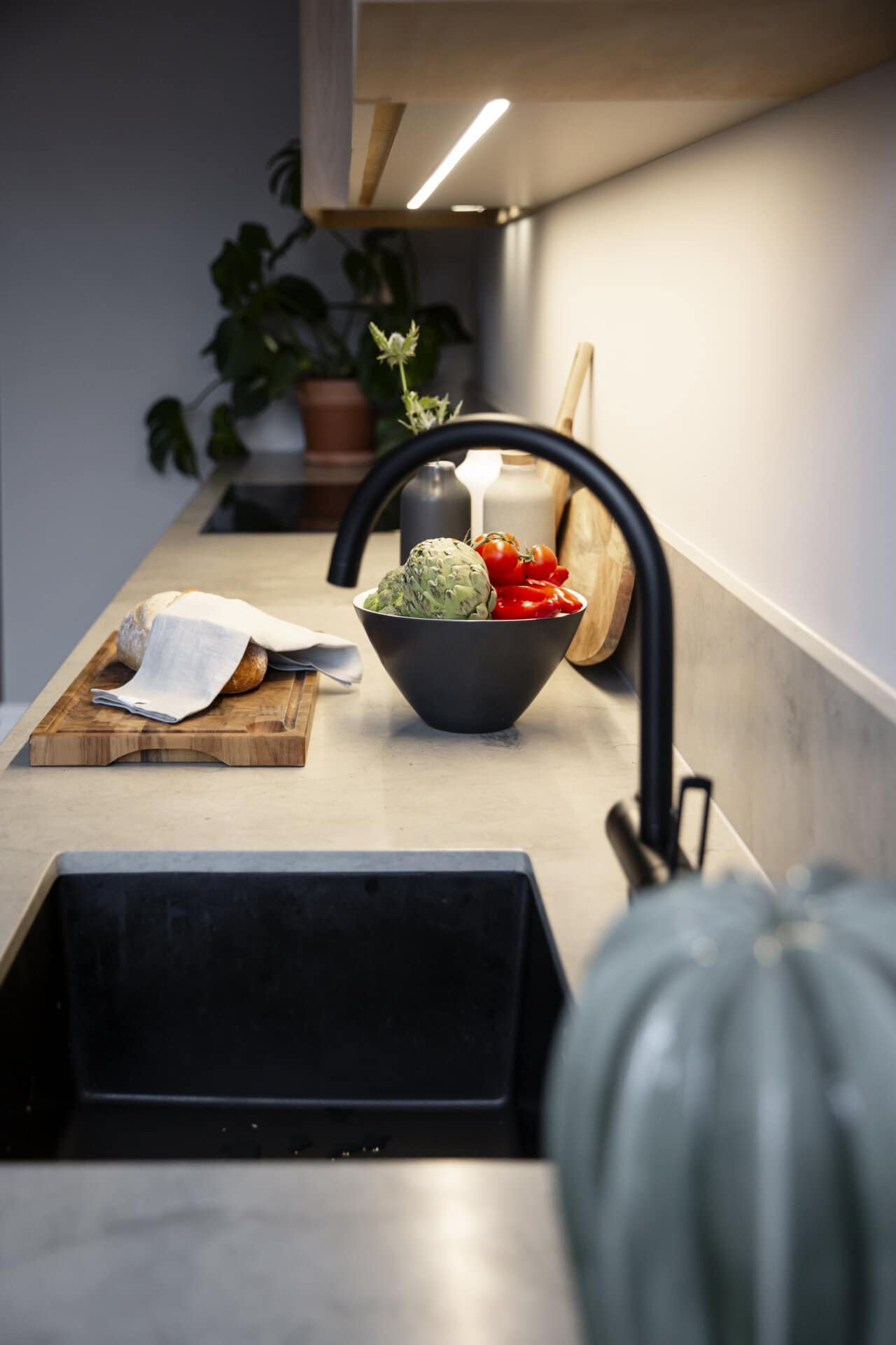 Stiligt - Modern köksbänk med en stiligt svart diskho, en skärbräda i trä med färskt bröd, en skål med färgglada grönsaker och en frodig växt i bakgrunden.