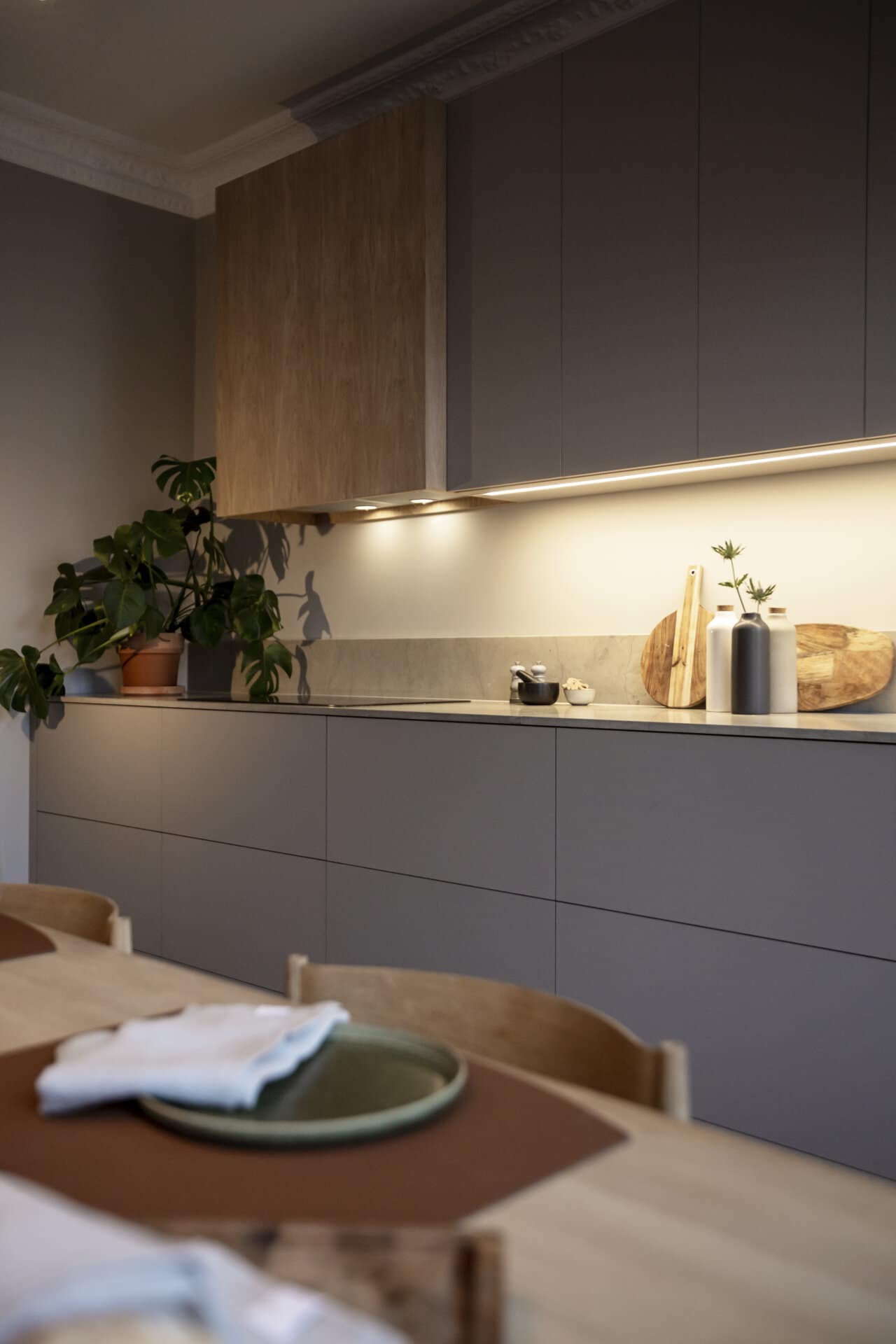 Stiligt - Modernt kök med stiligt gråa skåp, belysning under skåpet och en bänkskiva i trä. En växt och skärbrädor finns utställda. Ett bord med bordstabletter och servetter pryder förgrunden, vilket bidrar till den chica estetiken.