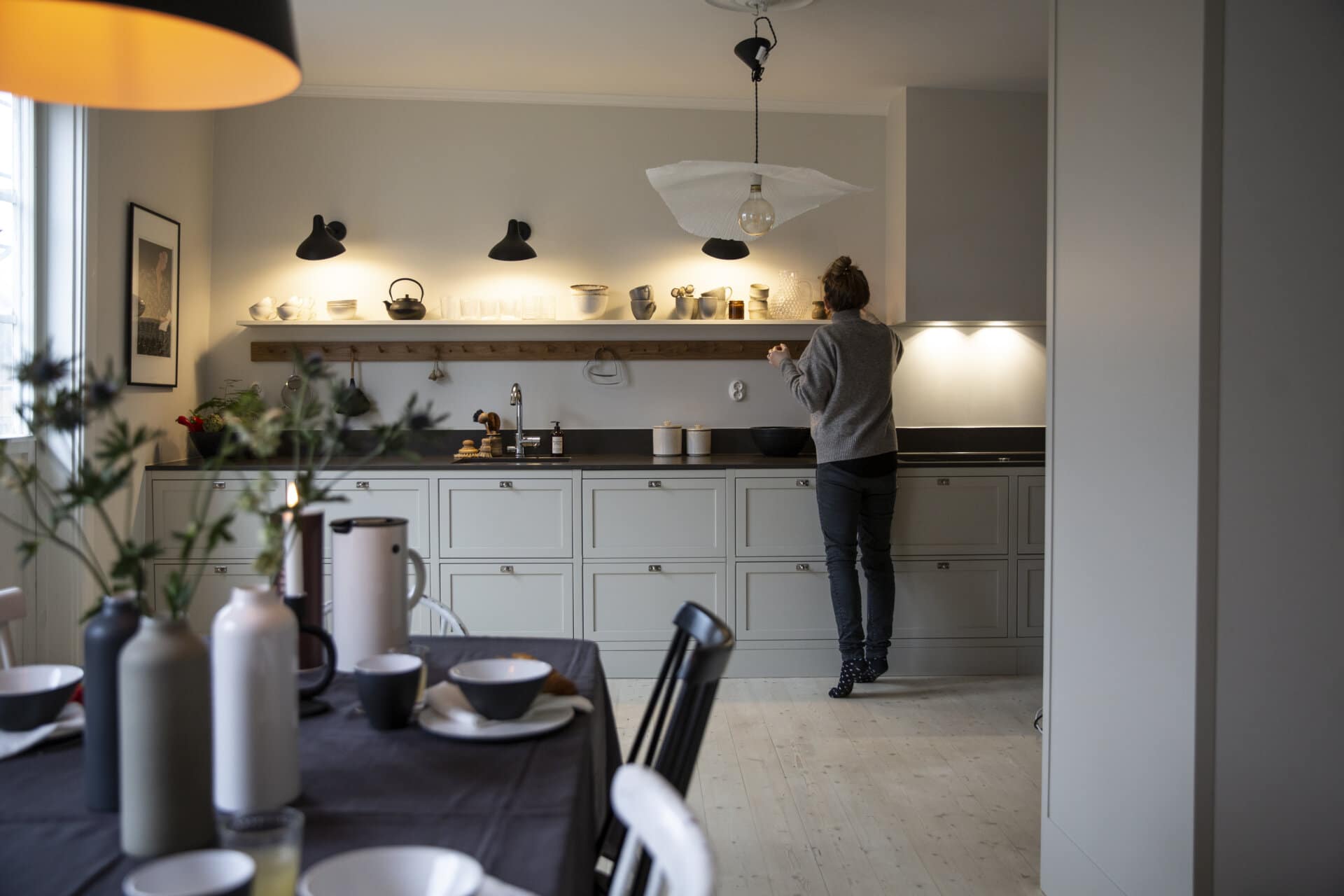 Stiligt - En person står i ett modernt kök med stiligt ljusa skåp och justerar en kopp på hyllan. I förgrunden finns ett elegant bord med eleganta fat och vaser.