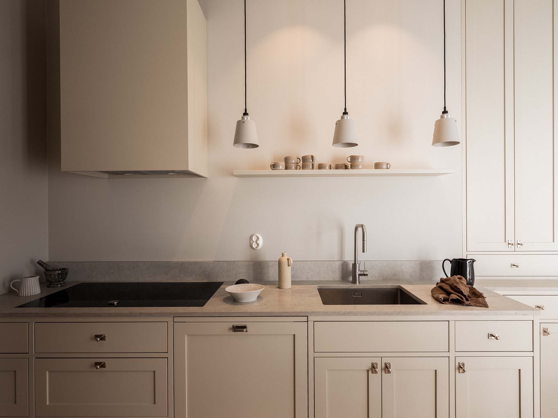 Stiligt - Ett stiligt minimalistiskt kök har beige skåp, spishäll, diskbänk och taklampor. En hylla visar snyggt staplade koppar och tallrikar. På bänkskivan hittar du en handduk och en vattenkokare som ger både funktion och stil.