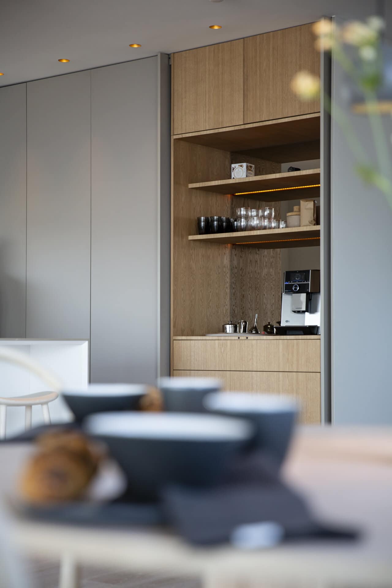 Stiligt - Det stiligt minimalistiska köket har ljusa träskåp och en inbyggd kaffebryggare. Hyllor rymmer glas och koppar, medan en del av ett matbord i förgrunden visar upp skålar och ett bakverk.