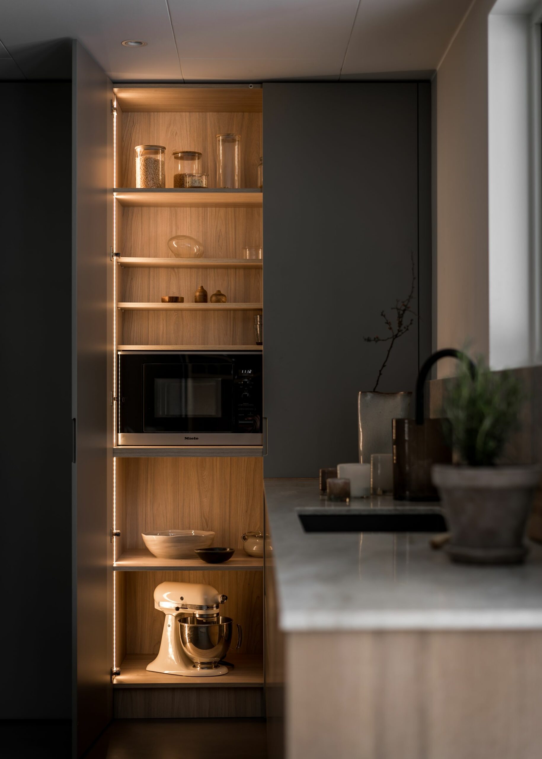 Stiligt - Ett modernt skafferi med stiligt inbyggda hyllor rymmer en mikrovågsugn, burkar, glas, en ställmixer och skålar. Mjuk belysning lyser upp träinredningen. En snygg bänkskiva syns i förgrunden.