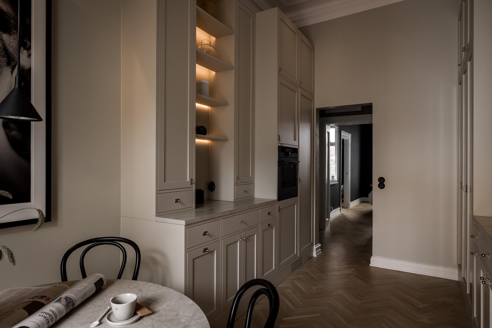 Stiligt - Ett svagt upplyst kök med stiligt beiga skåp, ett marmorbord med en kopp och en tidning. En hall med fiskbensgolv leder stilfullt till ett annat rum.