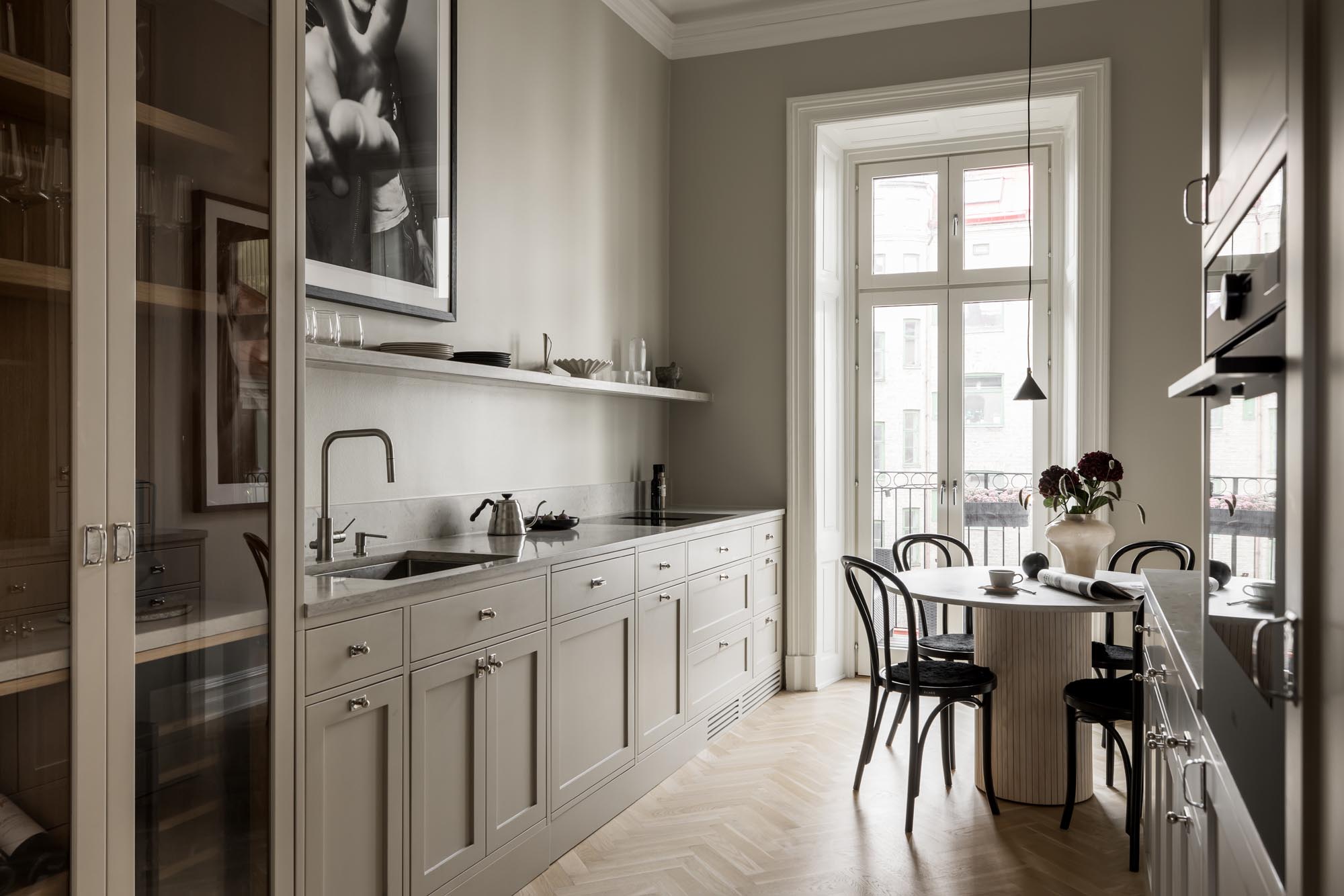 Stiligt - Det minimalistiska köket ståtar med stiligt ljusgrå skåp, en lång bänkskiva och ett litet matbord med fyra svarta stolar vid ett stort fönster. Monokromt konstverk hänger elegant på väggen.