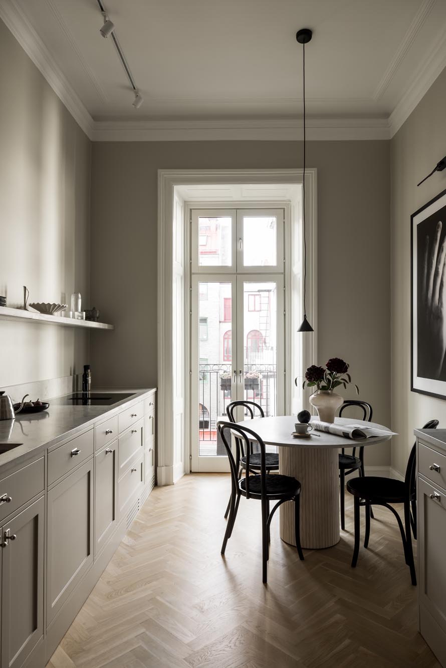 Stiligt - Ett stiligt minimalistiskt kök har ett runt matbord, svarta stolar och neutrala skåp. Ett stort fönster erbjuder balkongutsikt, kompletterat med vertikala konstverk på höger vägg.