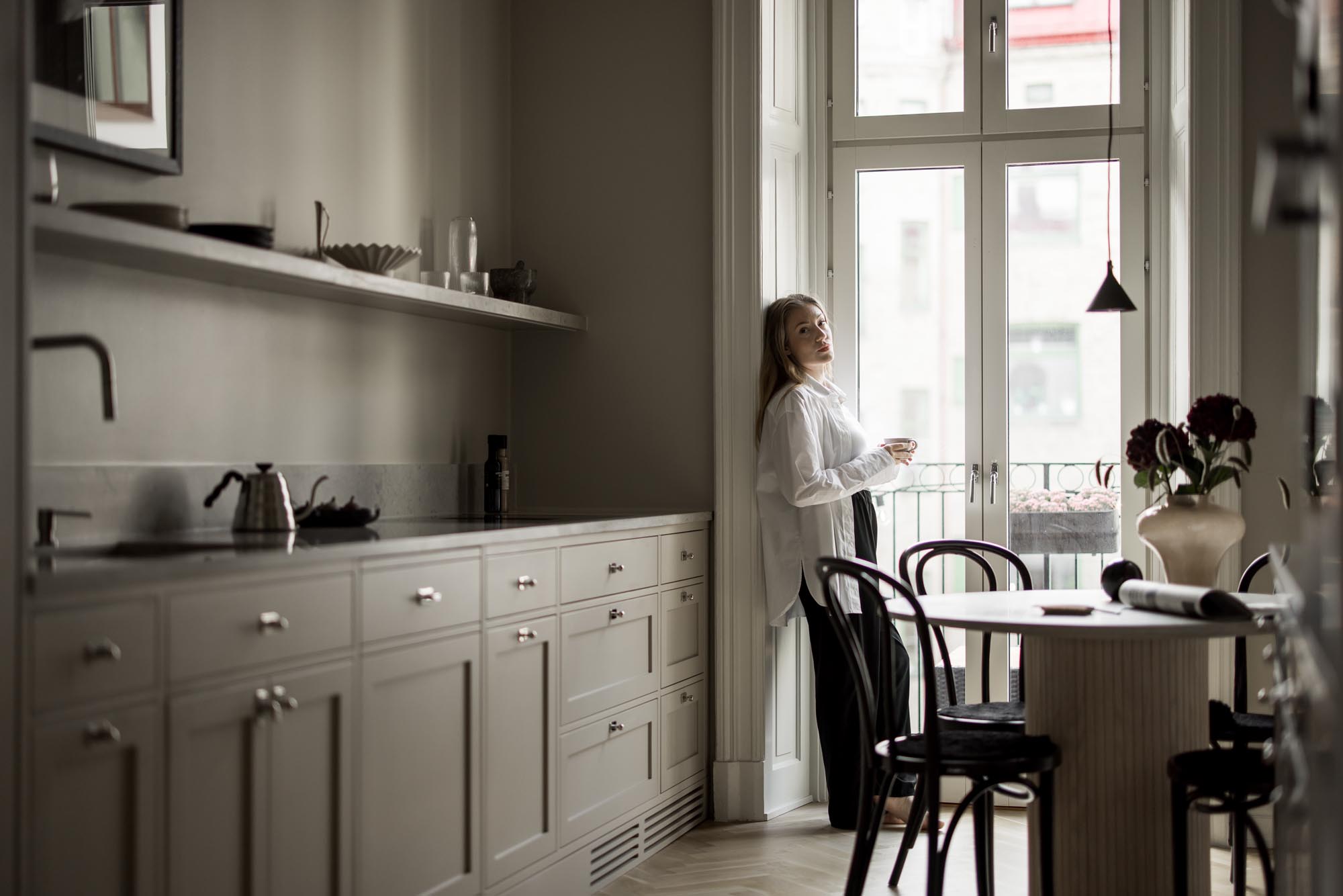 Stiligt - En person står i ett modernt stiligt kök med en kopp och stirrar ut genom ett stort fönster. Rummet ståtar med ljusa skåp och ett stilrent matbord med stolar.