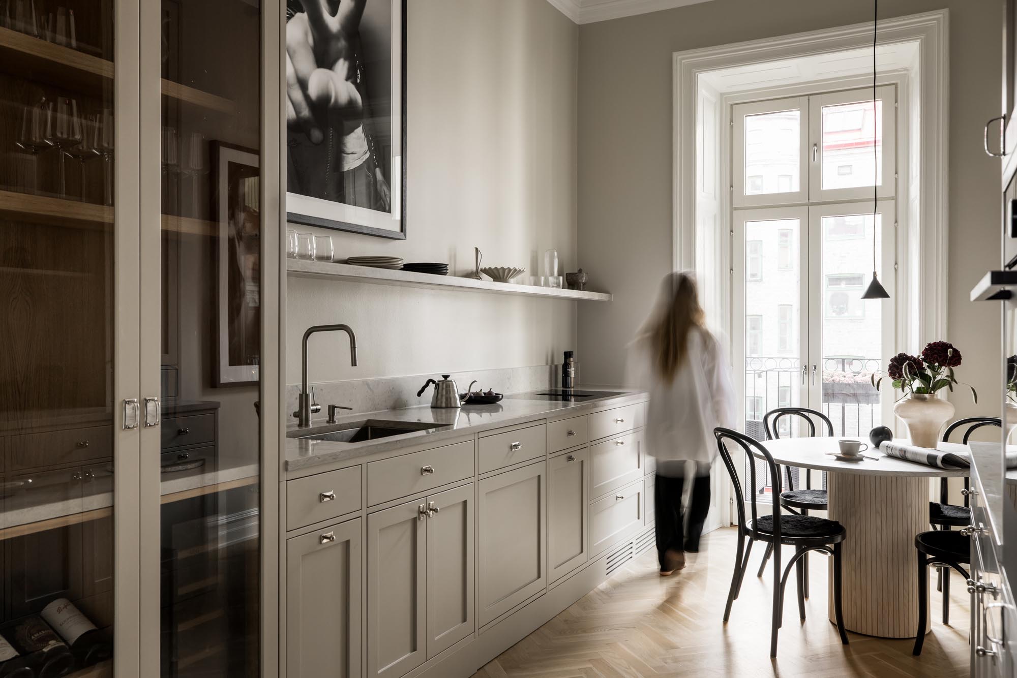 Stiligt - Ett Kök i shakerstilt som har beige skåp, en elegant svart bänkskiva och öppna hyllor. En person strosar förbi matbordet vid ett stort fönster, där inramade konstverk pryder väggen.