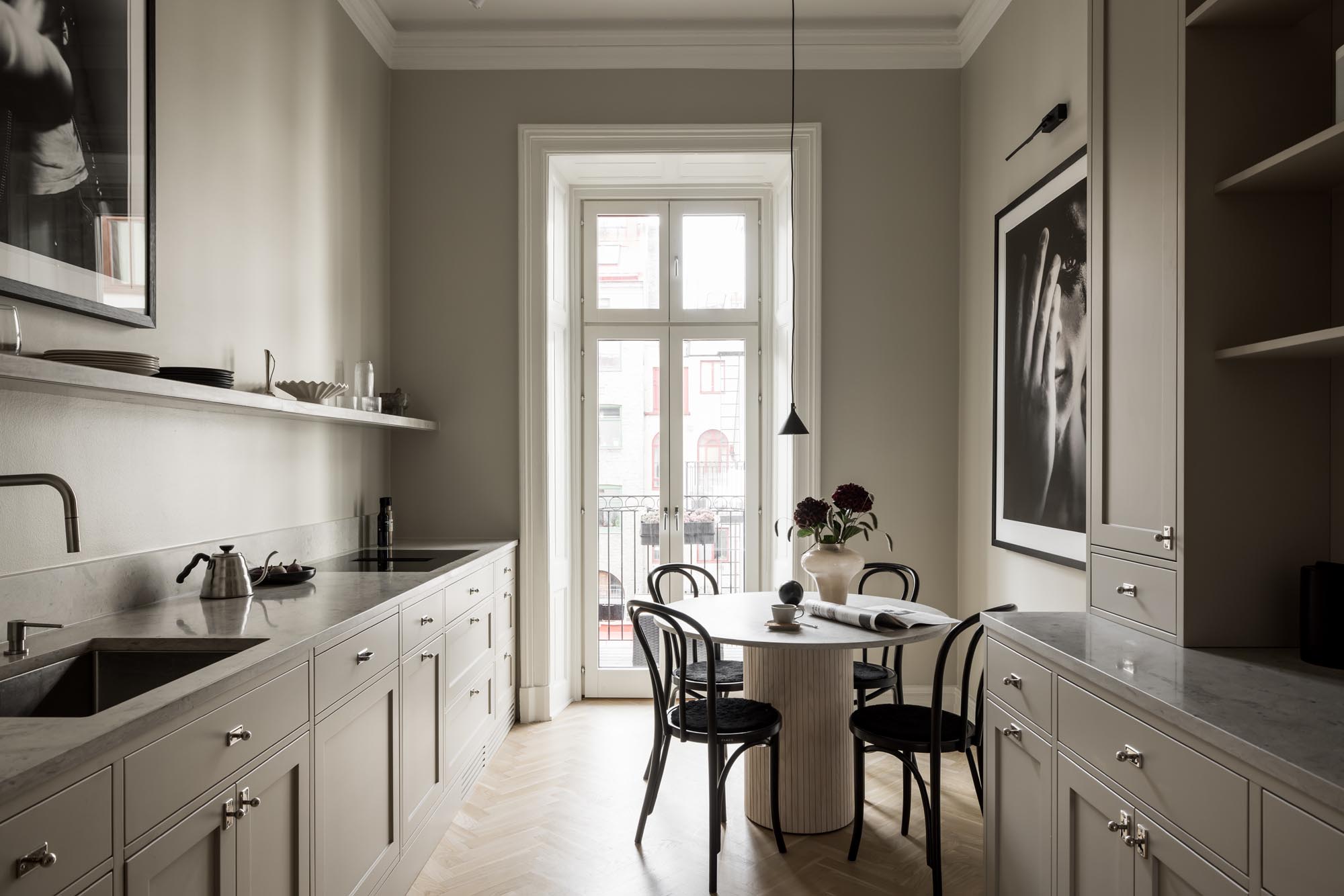 Stiligt - Minimalistiskt kök med ljusgrå skåp, ett runt matbord och svarta stolar som utstrålar en stiligt vibe. Stort fönster erbjuder utsikt över byggnader. Konst pryder väggarna, kompletterat med en taklampa ovanför bordet.