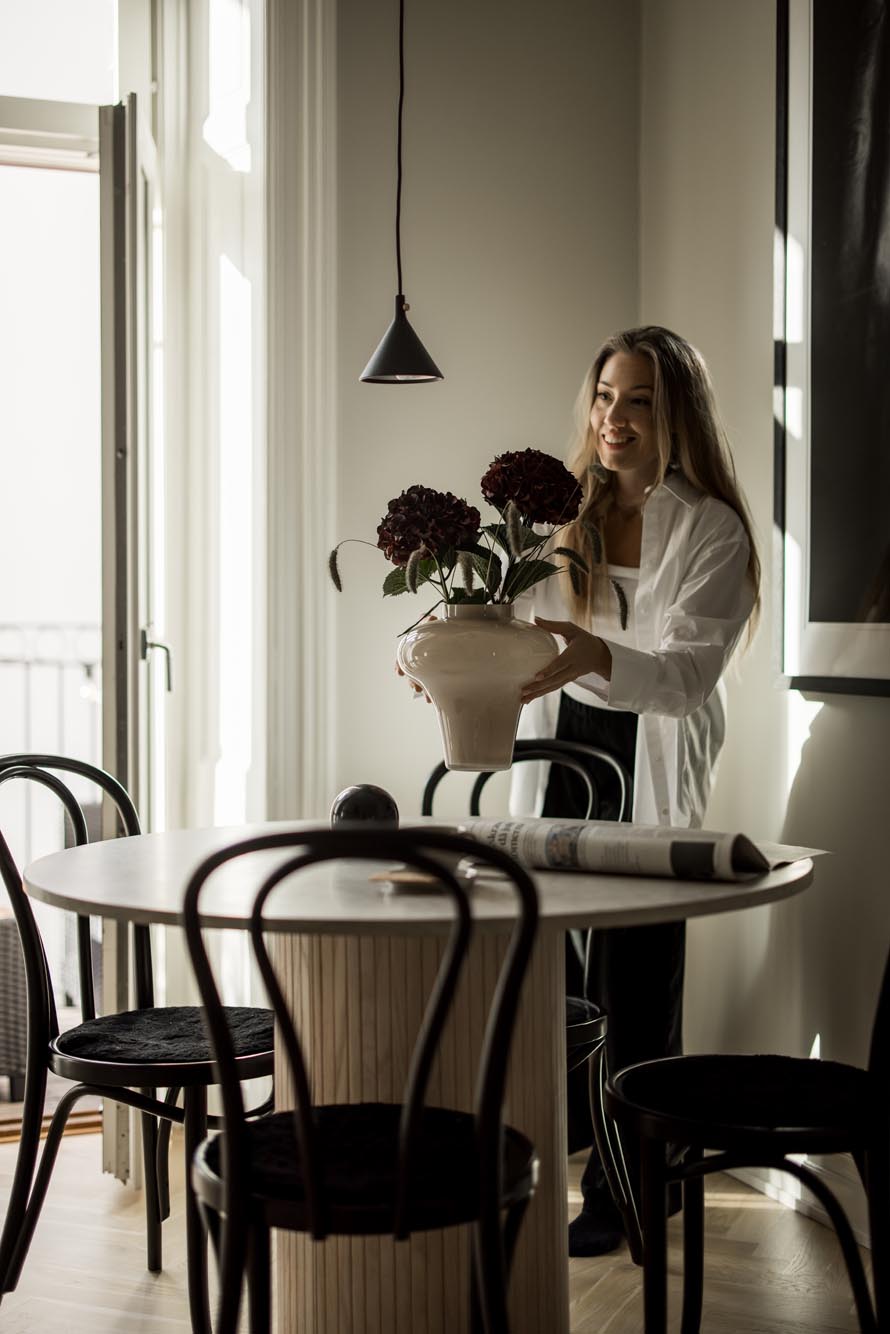 Stiligt - En person placerar en krukväxt på ett stiligt runt matbord i ett rum med svarta stolar och en hänglampa.