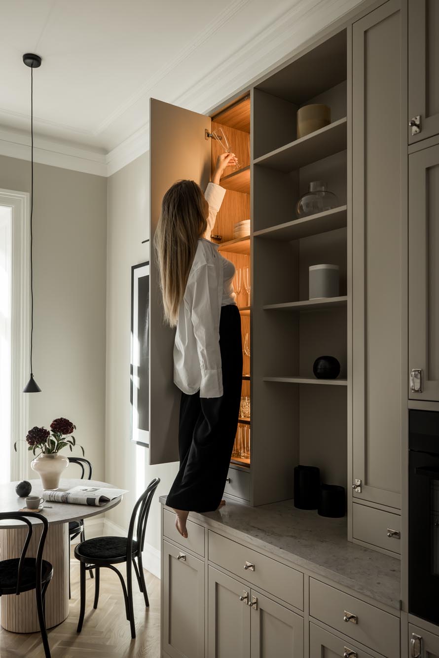 Stiligt - En person som står på en snygg köksbänk sträcker sig in i ett högt stiligt skåp. Rummet har ett elegant bord, stolar och taklampa.