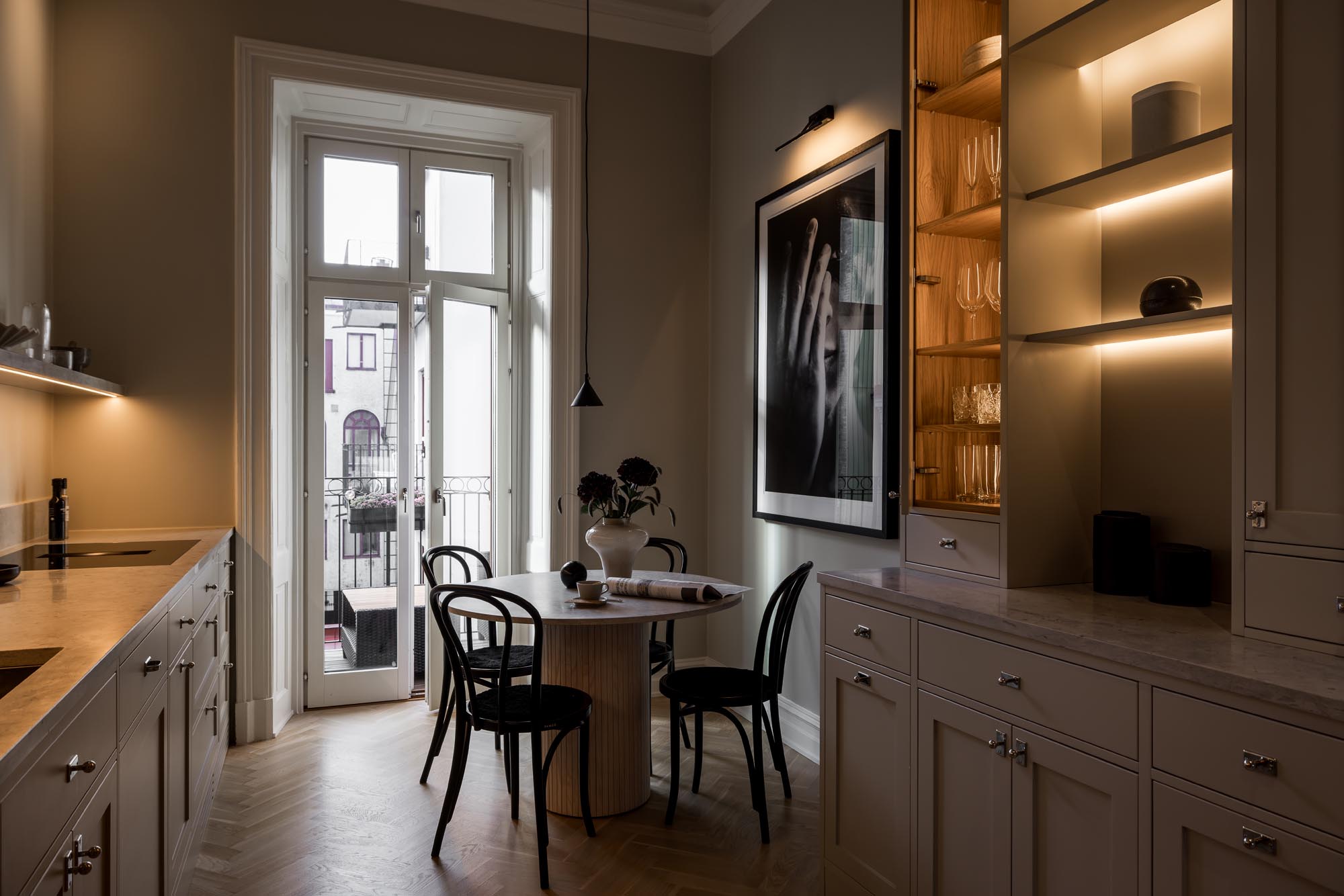 Stiligt - Ett modernt kök med stiligt ljusskåp, ett runt matbord med stolar och ett stort fönster som leder till en balkong. Det finns ett svartvitt foto på väggen och mjuk omgivningsbelysning.