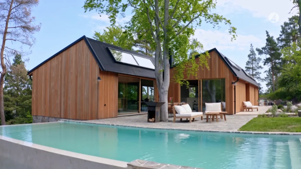 Stiligt - Ett modernt trähus med stora glasfönster ligger bredvid en rektangulär utomhuspool, med utemöbler och ett träd på terrassen.