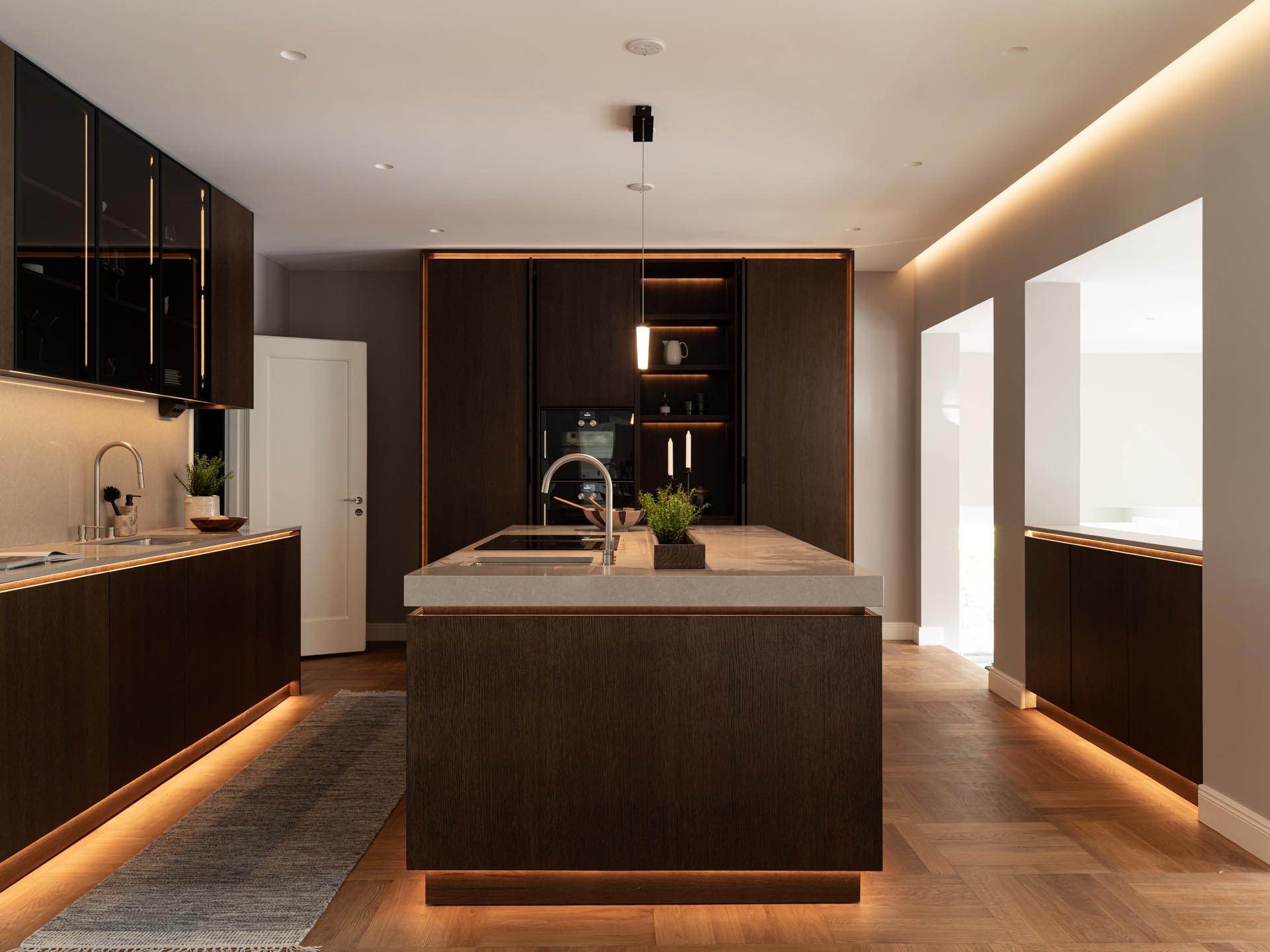 Stiligt - Modern träkök med pocketdörrar, mörka träskåp, central köksö med vask, under-skåpsbelysning, integrerad vitvaror och minimalistisk inredning.
